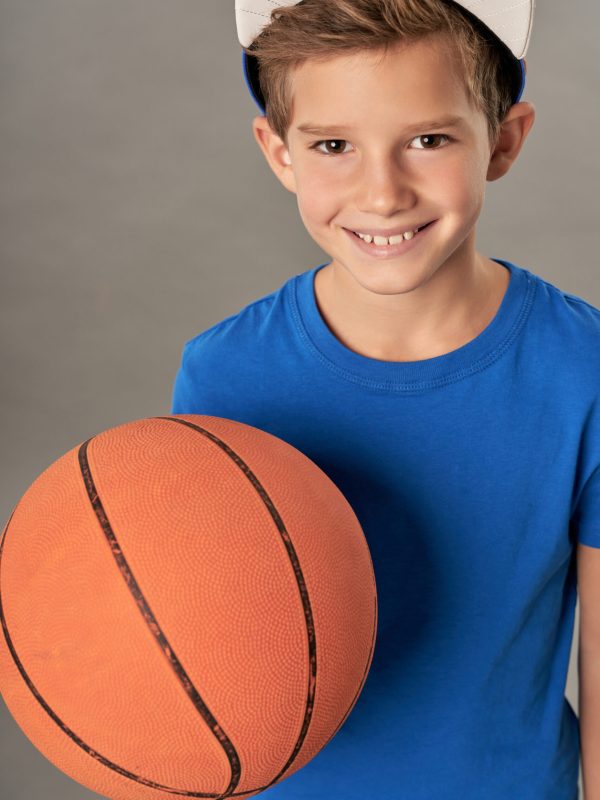 Adorable boy basketball player holding game ball and smiling while standing against gray background